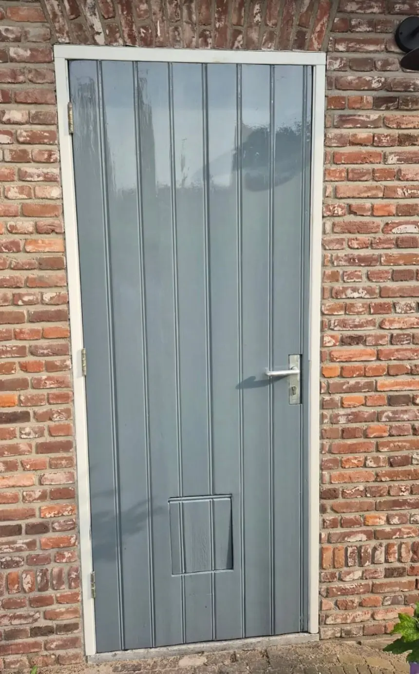 Grijze buitendeur met verticale panelen en wit kozijn in een bakstenen muur