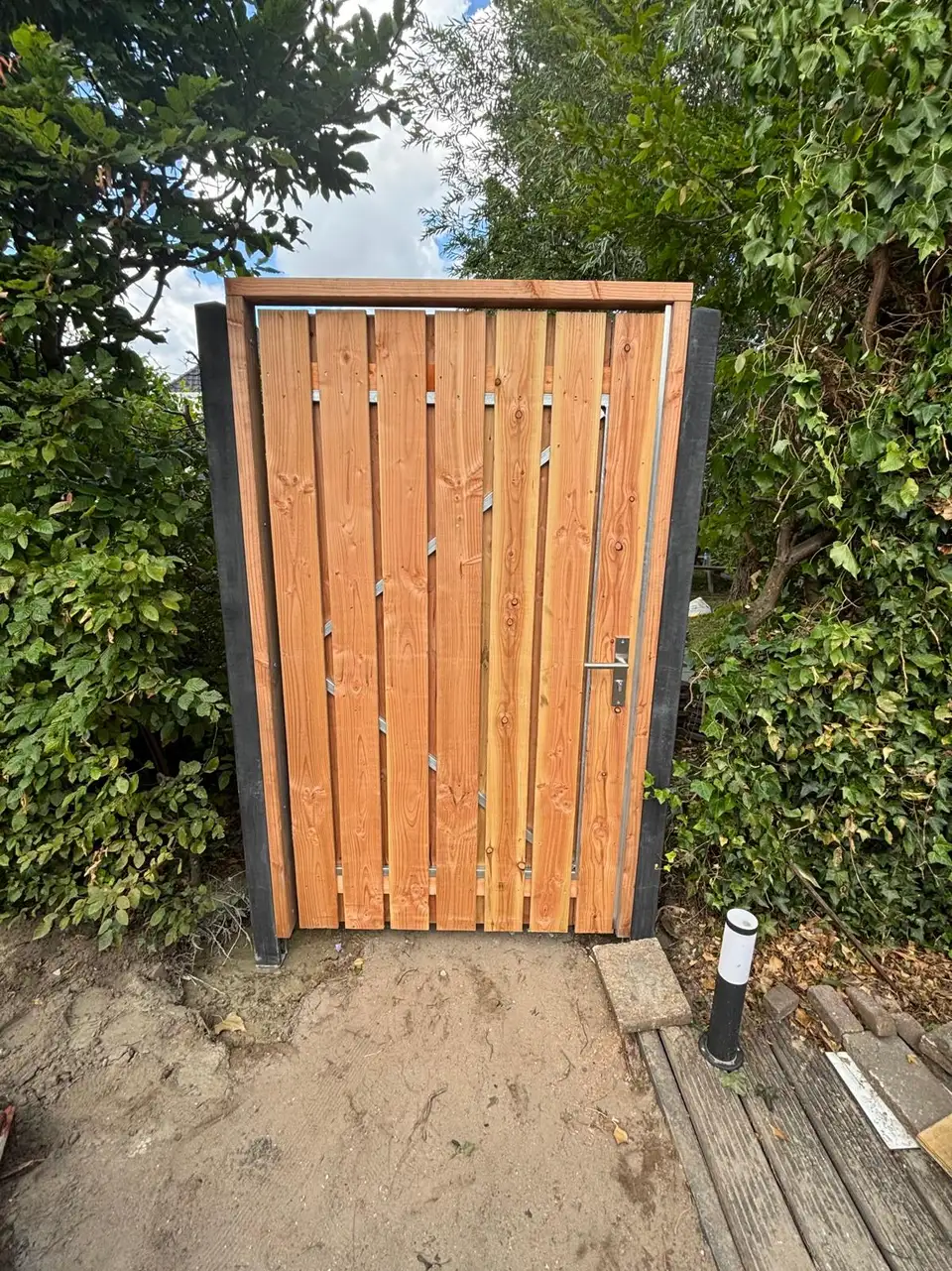 Nieuwe houten tuinpoort met deurkruk tussen twee zwarte palen, omgeven door groen