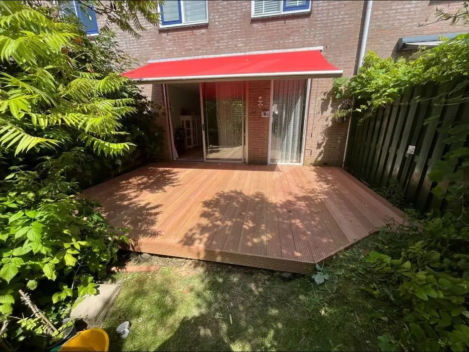 Nieuw houten vlonderterras achter een rijtjeshuis met een rood zonnescherm en groene borders